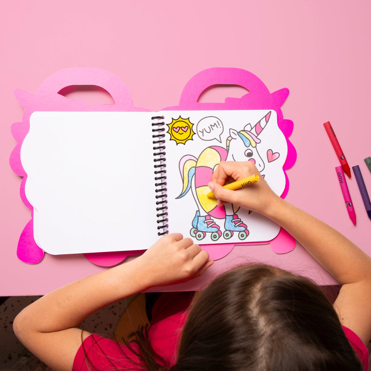 Child coloring a unicorn on a pink table with coloring book and markers.
