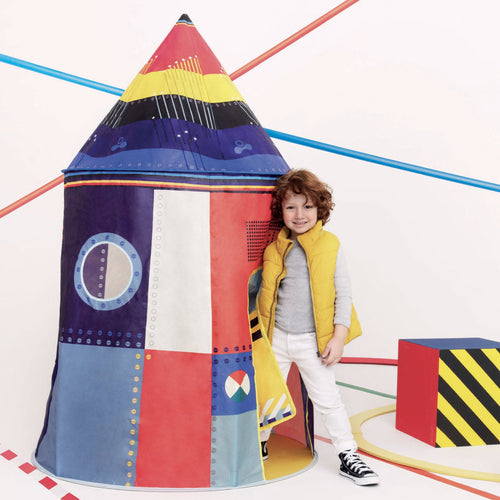 Child standing next to a colorful rocket-shaped playhouse on a white background