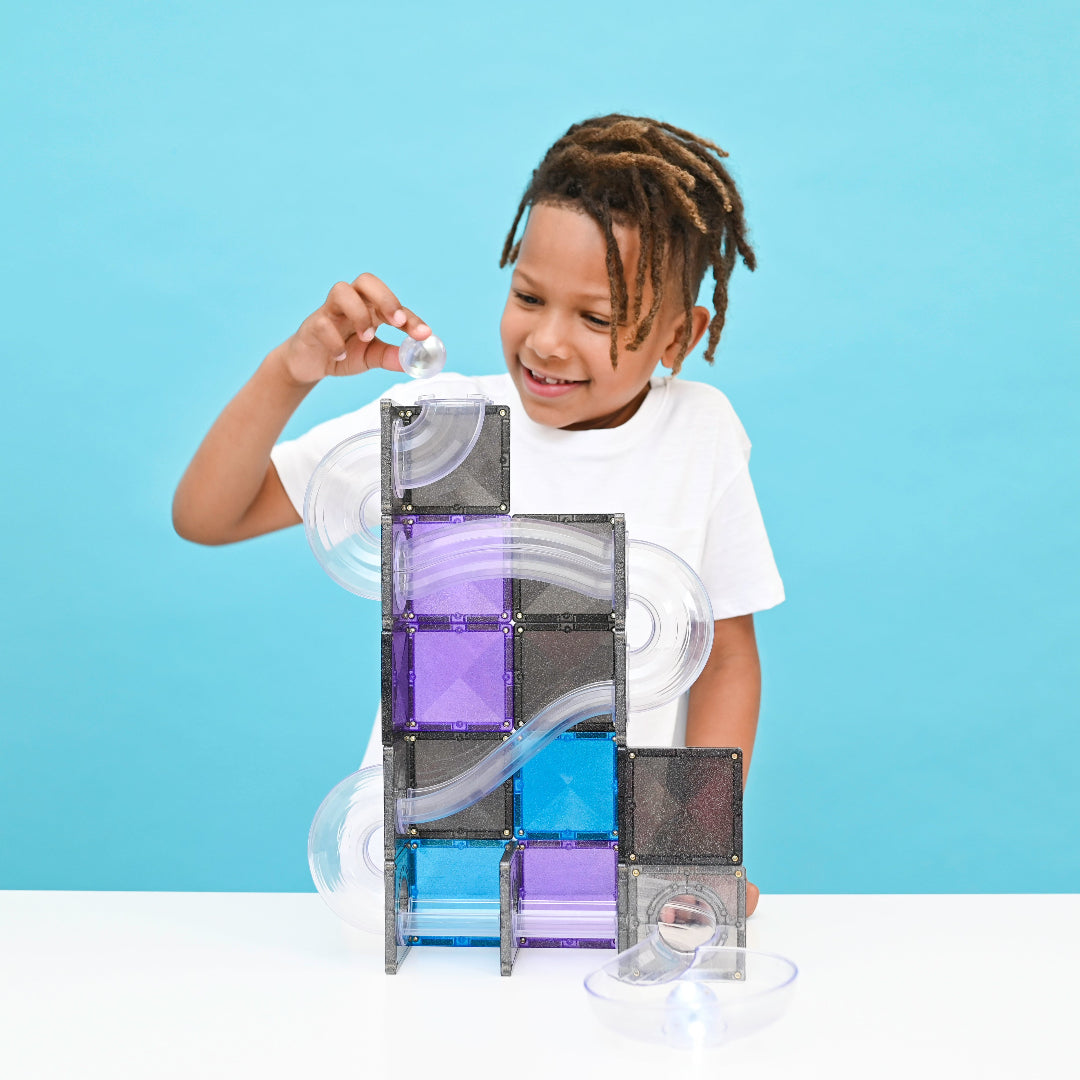 Smiling boy building a CONNETIX Ball Run Galaxy tower on a bright blue background
