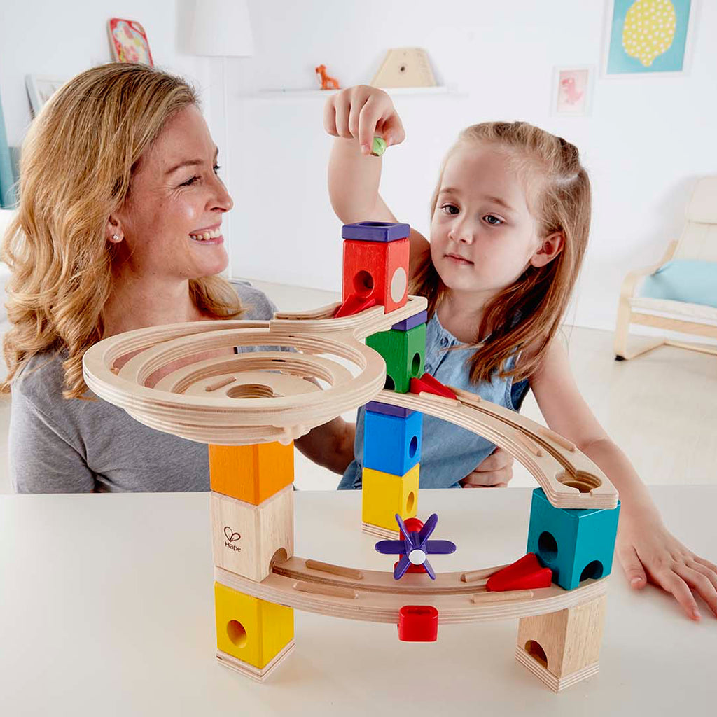 Woman and child playing with a colorful wooden marble run toy in a bright living room.