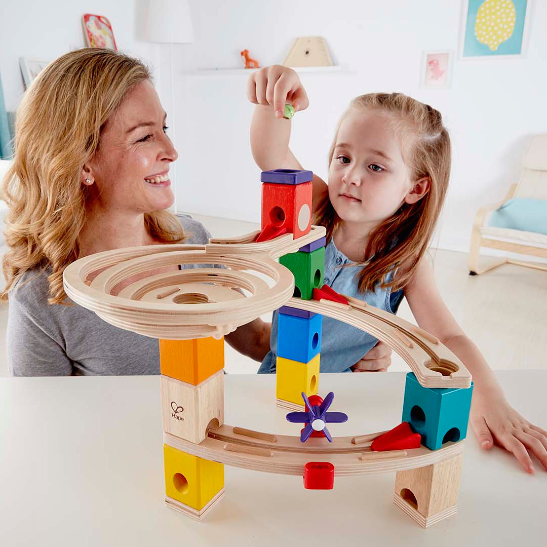 Woman and child playing with a colorful wooden marble run toy in a bright living room.