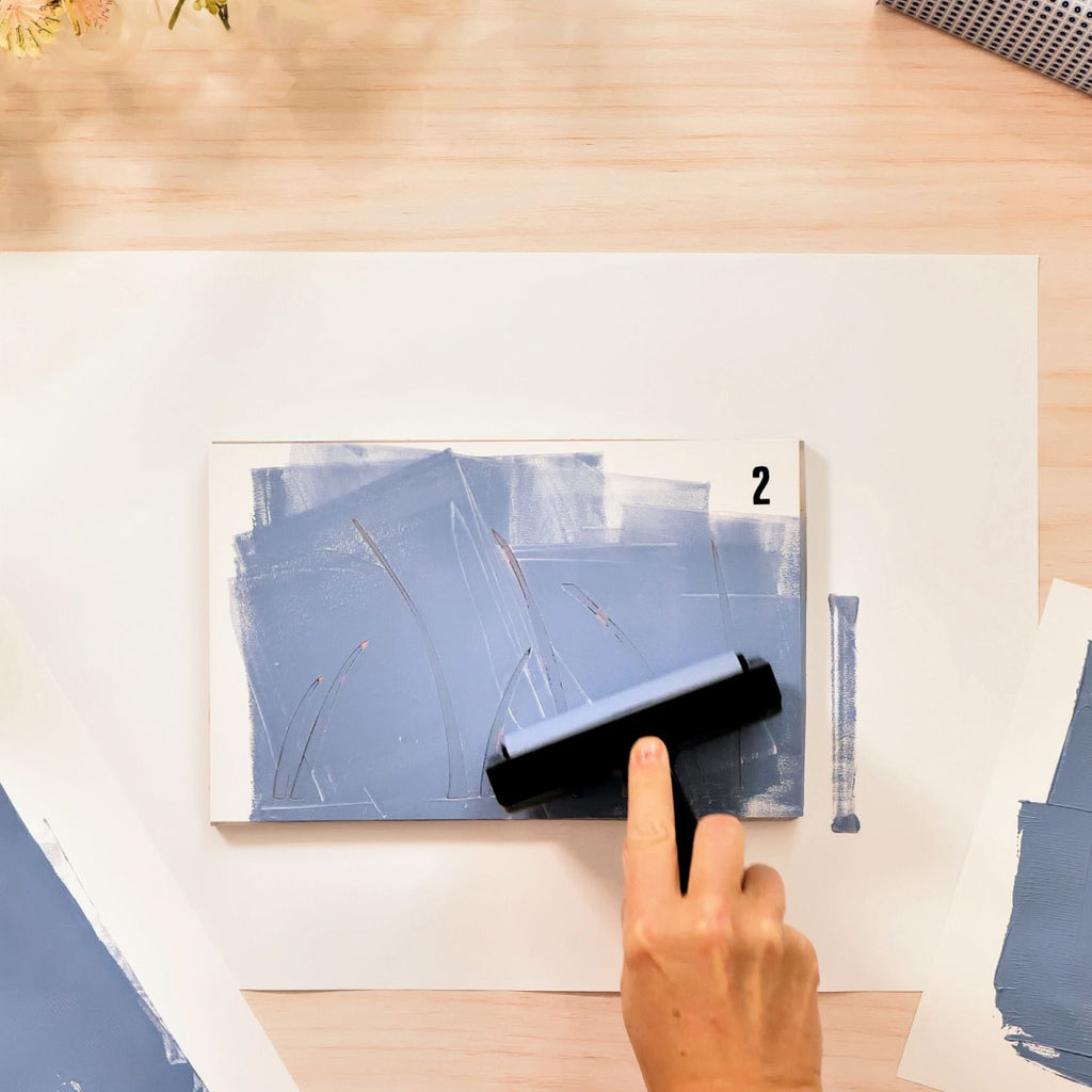 Person using a brayer on a piece of paper with blue abstract design on a wooden table.