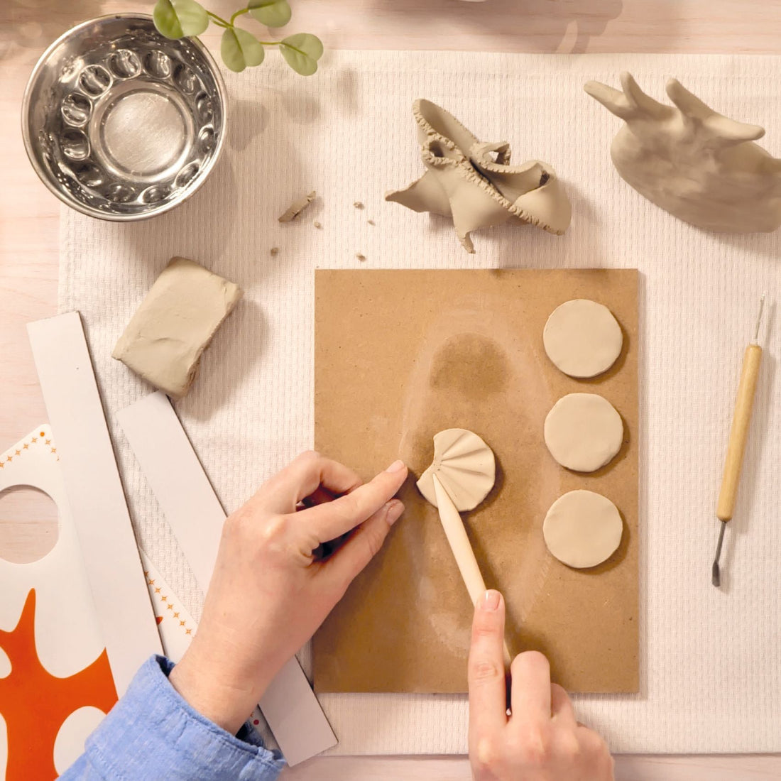 Person working with clay on a wooden board, surrounded by various clay items and tools on a light surface.
