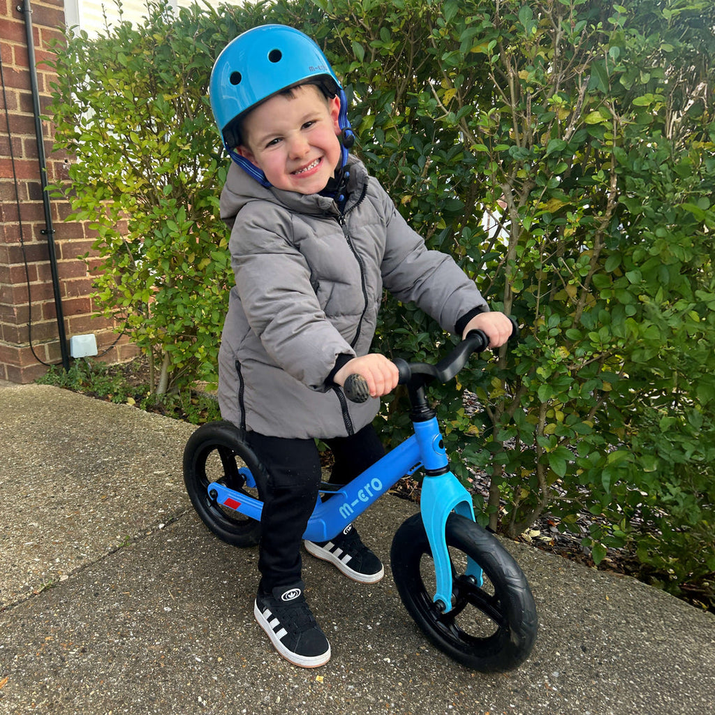 Smiling child in a blue helmet and grey coat sitting on the blue Micro Balance Bike Lite