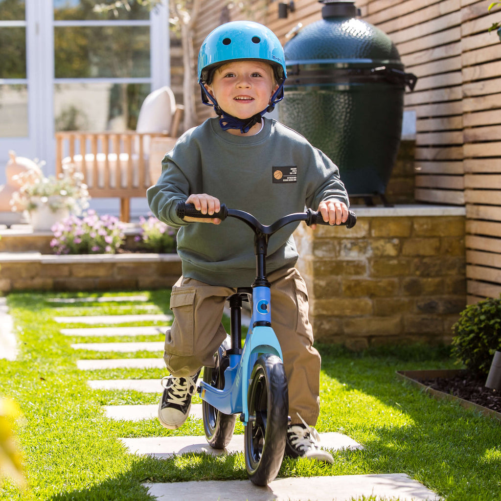 Smiling boy in a blue helmet riding the blue Micro Balance Bike Lite across a garden lawn