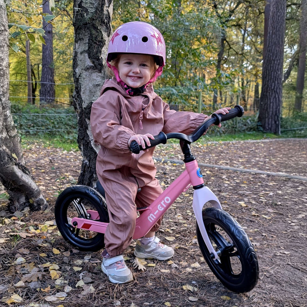 Smiling child in a lilac helmet and pink suit sitting on the pink Micro Balance Bike Lite in a forest