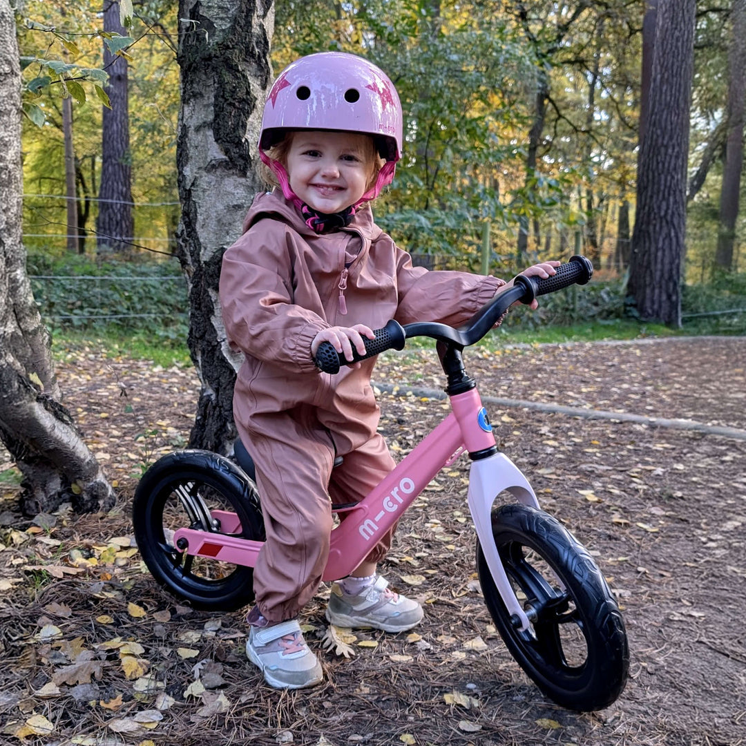 Smiling child in a lilac helmet and pink suit sitting on the pink Micro Balance Bike Lite in a forest