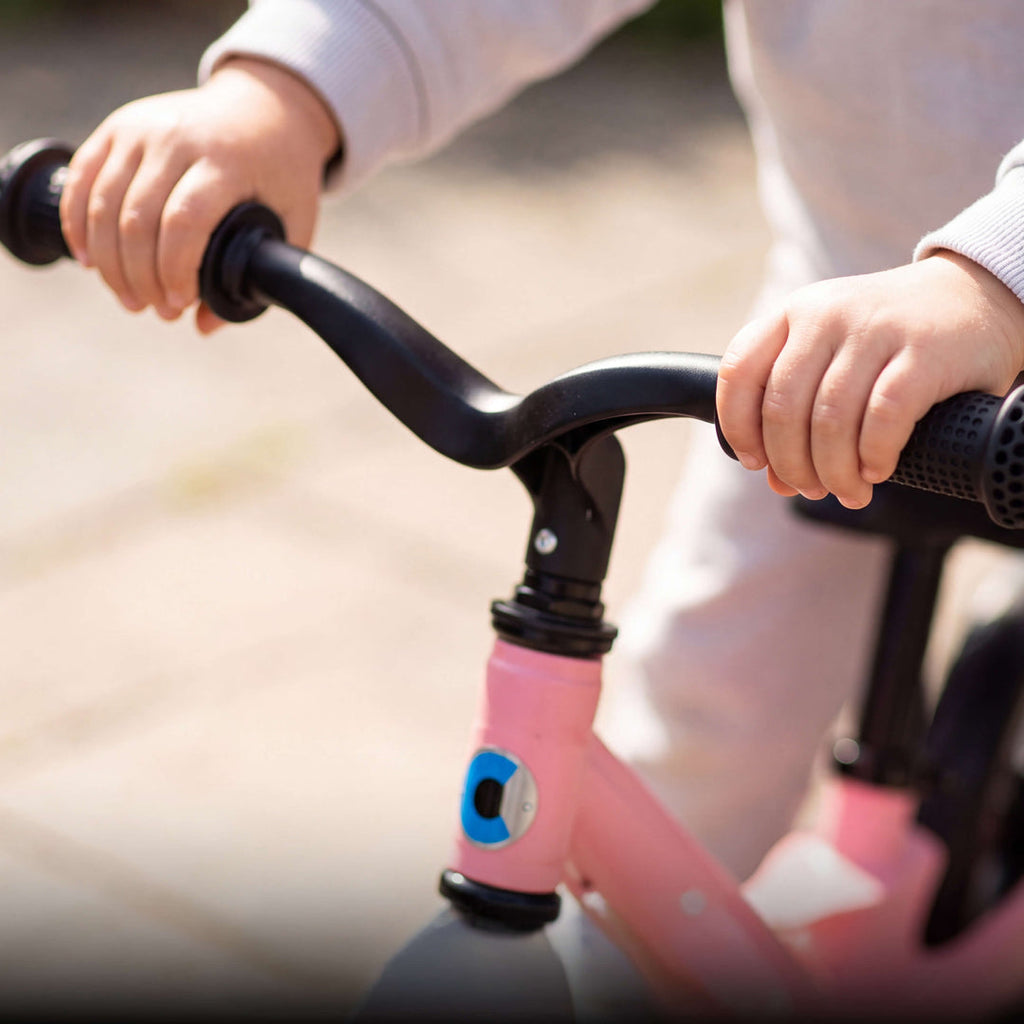 Close-up of a child's hands on the black handlebar grips of the pink Micro Balance Bike Lite