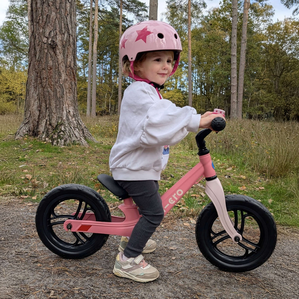 Child in a pink star helmet riding the pink Micro Balance Bike Lite through woodland