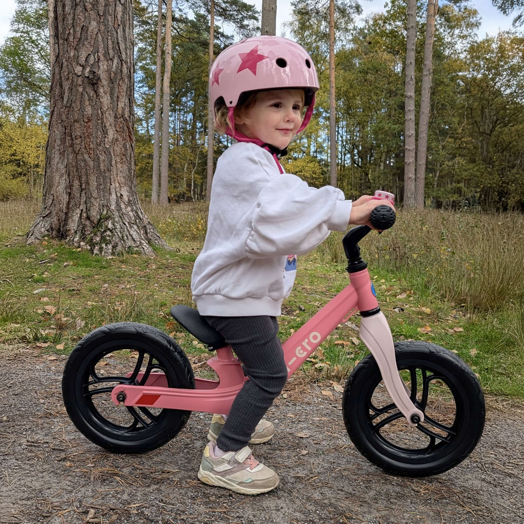 Child in a pink star helmet riding the pink Micro Balance Bike Lite through woodland