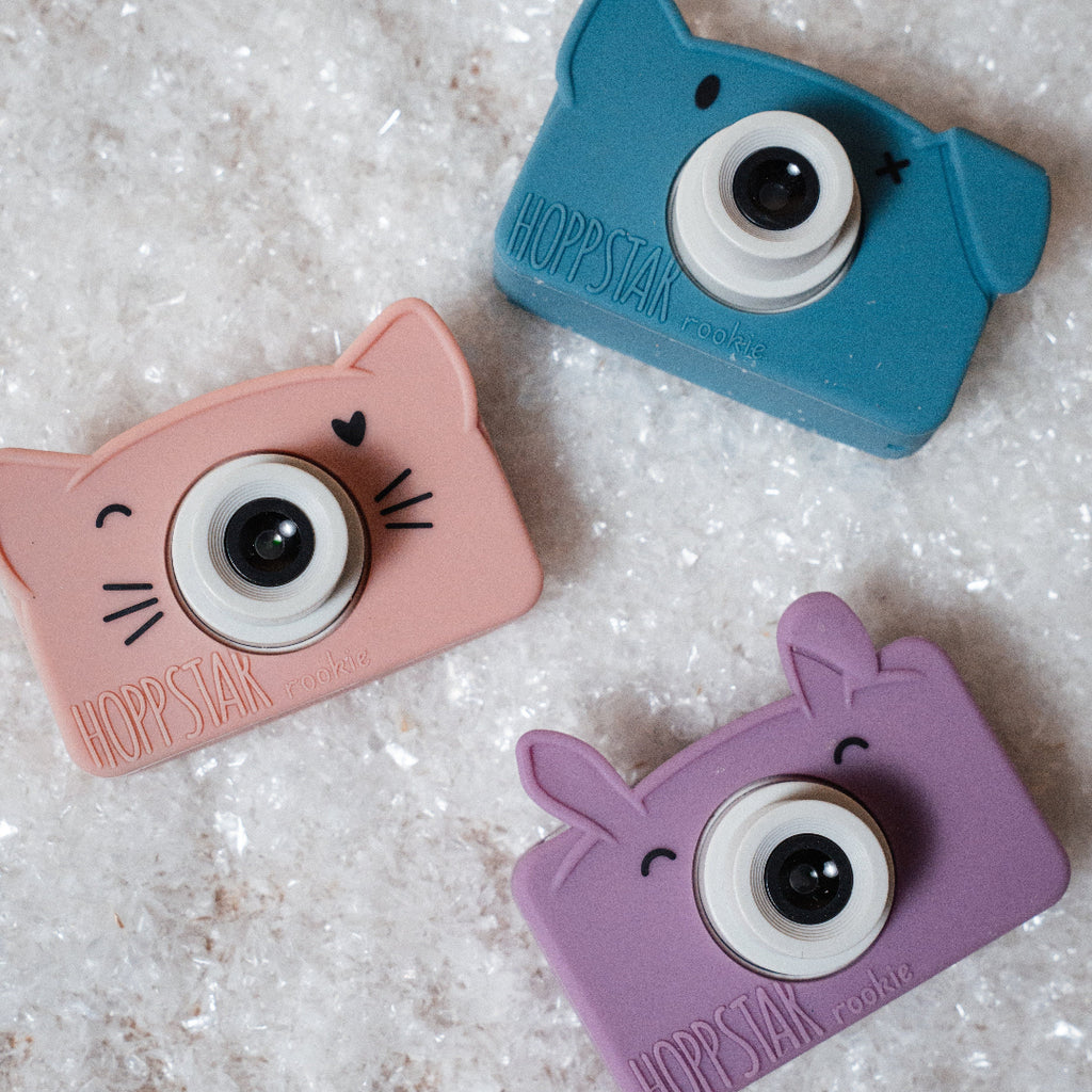 Three small cat-shaped camera accessories in pastel pink, blue, and purple, arranged on a textured white surface. Each has a large round black-and-white lens detail and the brand name HOPSTAR embossed near the bottom.
