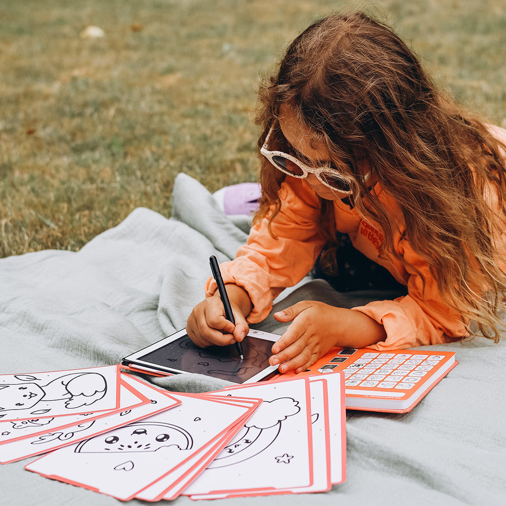 Child coloring outdoors on a blanket with coloring books and crayons.