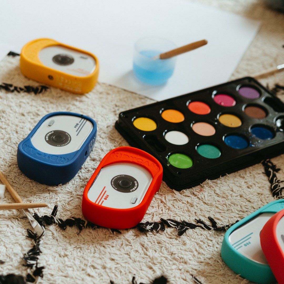 Orange, blue, red and turquoise KIDYCAM PRO cameras on a cream rug next to a watercolour paint palette