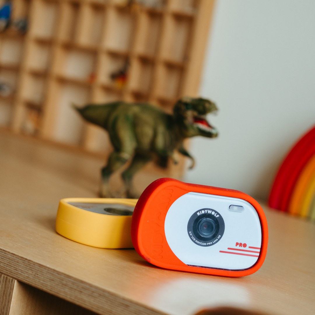 Red KIDYCAM PRO camera and an orange one lying beside it on a wooden desk, with a toy T-rex and wooden shelves behind