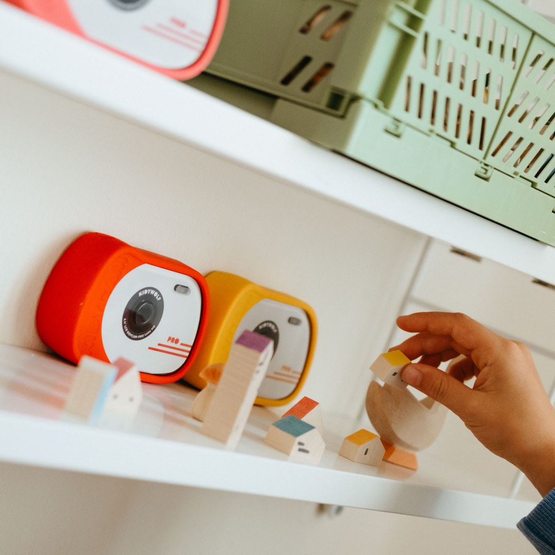 Red and orange KIDYCAM PRO cameras on a white shelf with wooden toy houses as a child's hand reaches in