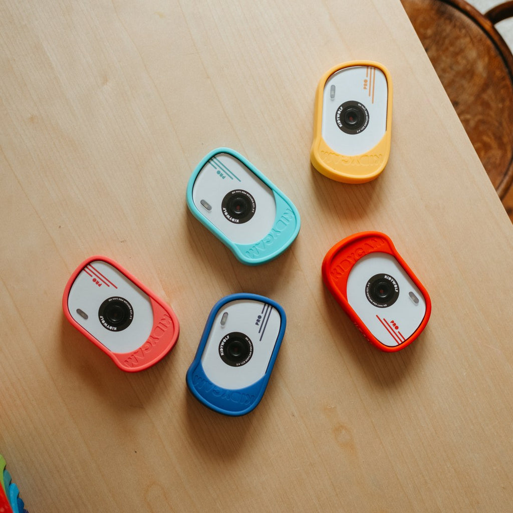 Five KIDYCAM PRO cameras in orange, turquoise, coral, blue and red laid out on a wooden table, seen from above
