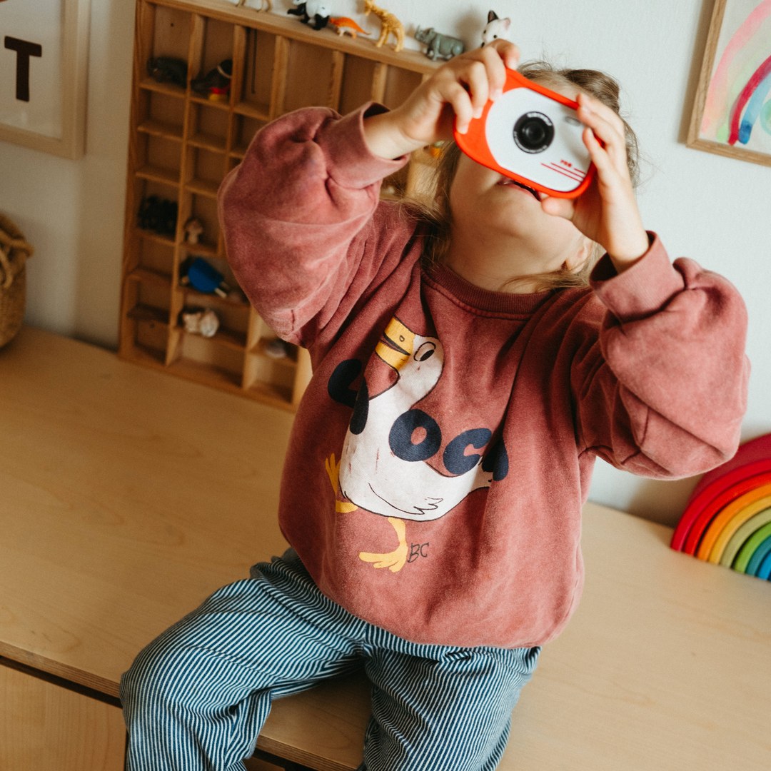 Toddler sitting on a desk holding a red KIDYCAM PRO up to their face to take a picture