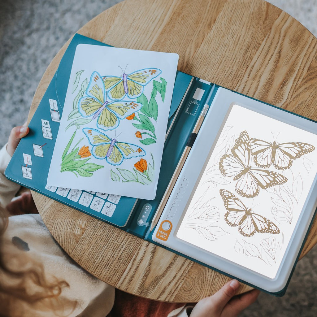KIDYDRAW Pad open on a round wooden table, the light pad glowing under a butterfly tracing card next to a finished coloured butterfly drawing
