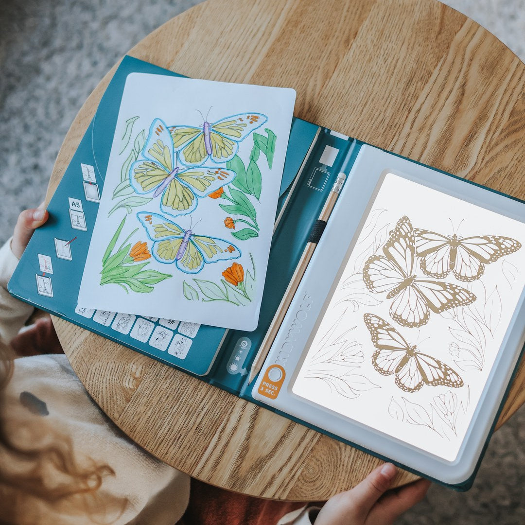 KIDYDRAW Pad open on a round wooden table, the light pad glowing under a butterfly tracing card next to a finished coloured butterfly drawing