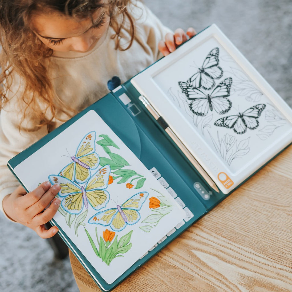 Curly-haired girl holding the open KIDYDRAW Pad, with a coloured butterfly drawing on one side and butterfly outlines on the light pad on the other