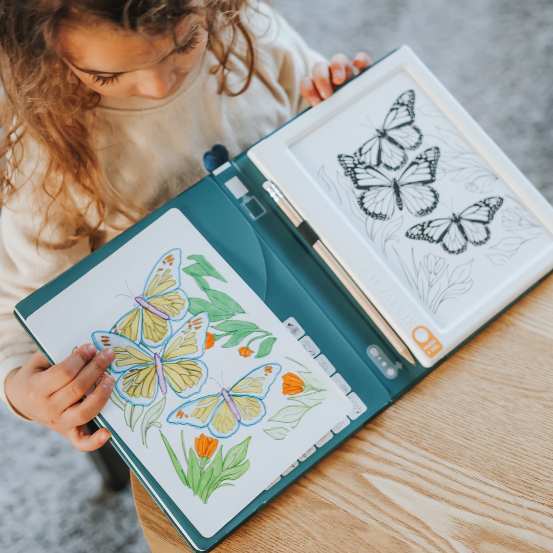 Curly-haired girl holding the open KIDYDRAW Pad, with a coloured butterfly drawing on one side and butterfly outlines on the light pad on the other