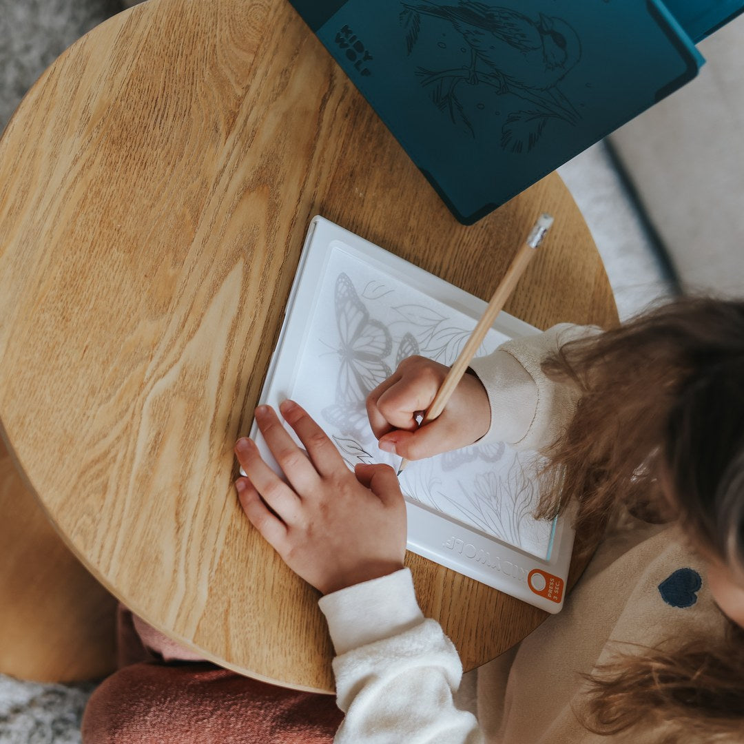 Child seen from above tracing a butterfly design with a wooden pencil on the KIDYDRAW light pad at a round wooden table