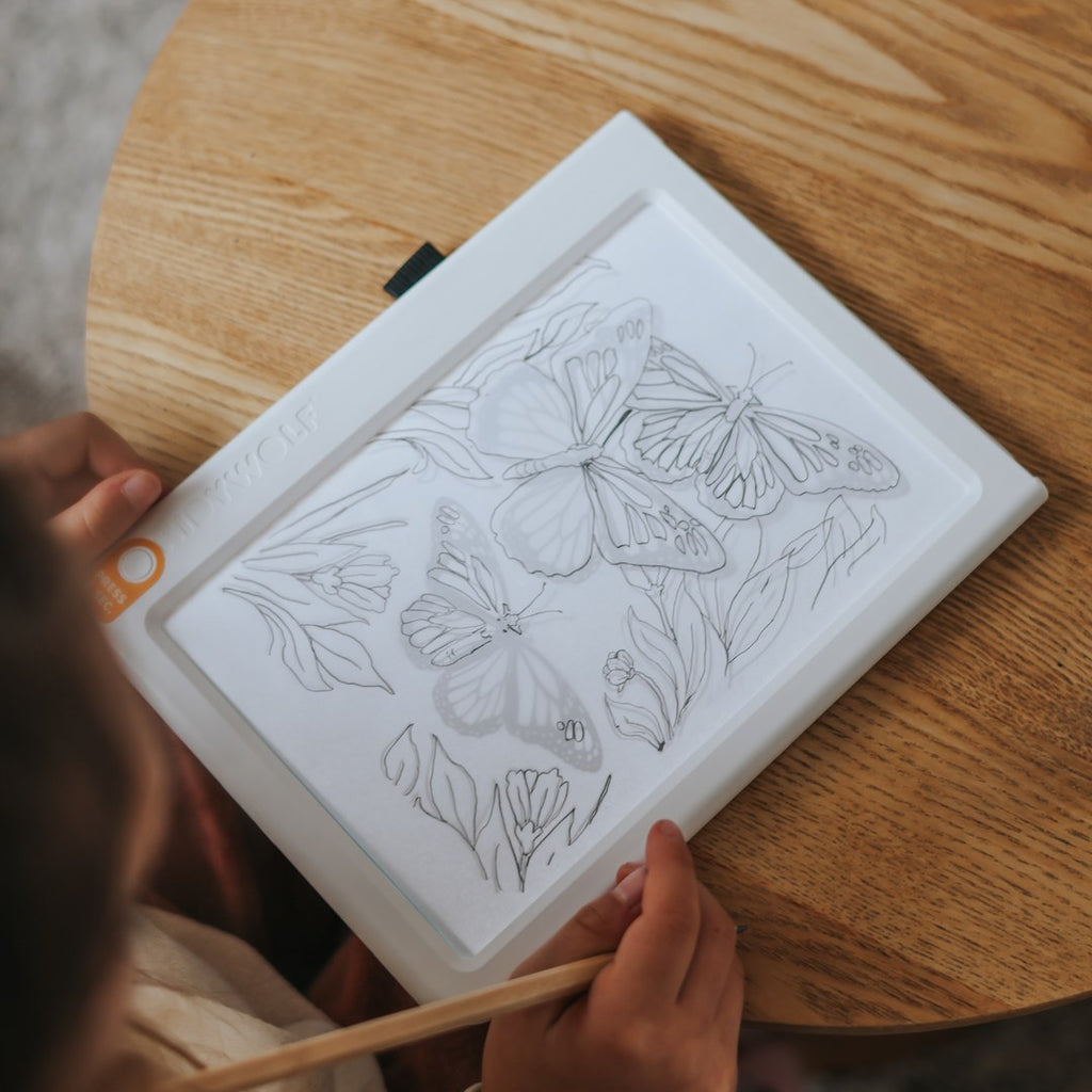 Close-up of a child's hands tracing butterflies and flowers in pencil on the KIDYDRAW light pad