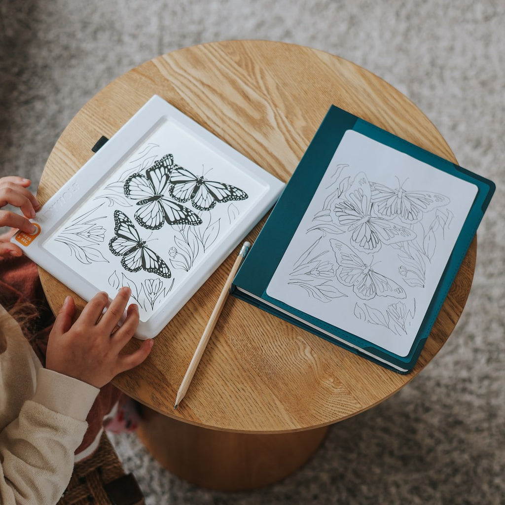 KIDYDRAW light pad with a butterfly tracing card, a pencil and the teal folder holding a traced drawing on a round wooden table
