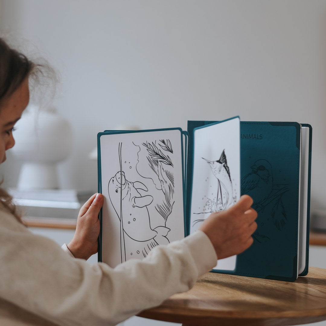 Child turning the tracing cards in the KIDYDRAW Pad at a wooden table, showing a sloth and a swordfish design