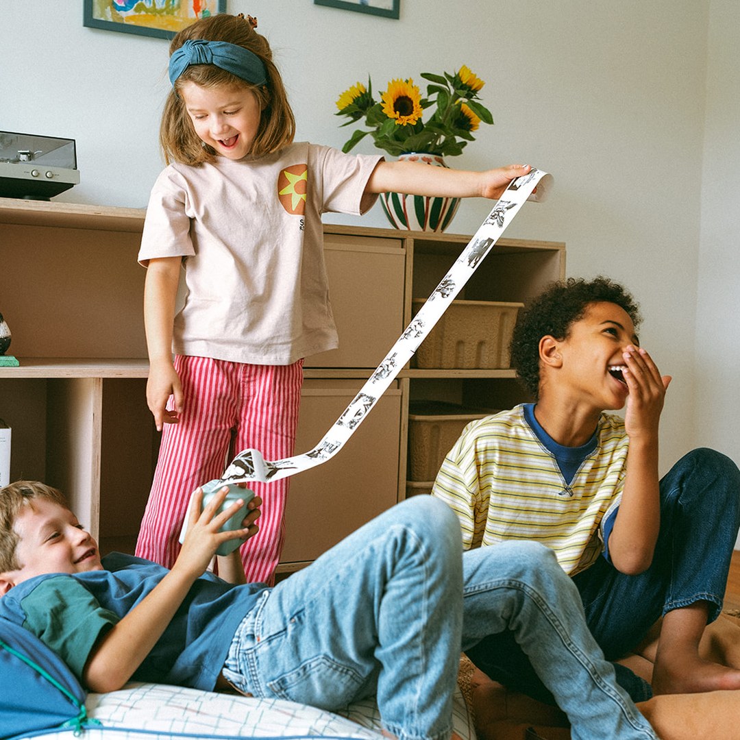 Girl standing on a sofa pulling a long printed strip from the green KIDYPRINTER AI while two boys laugh in a living room