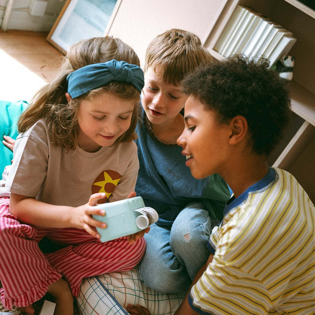 Three children huddled together watching a print come out of the green KIDYPRINTER AI held by a girl in a headband