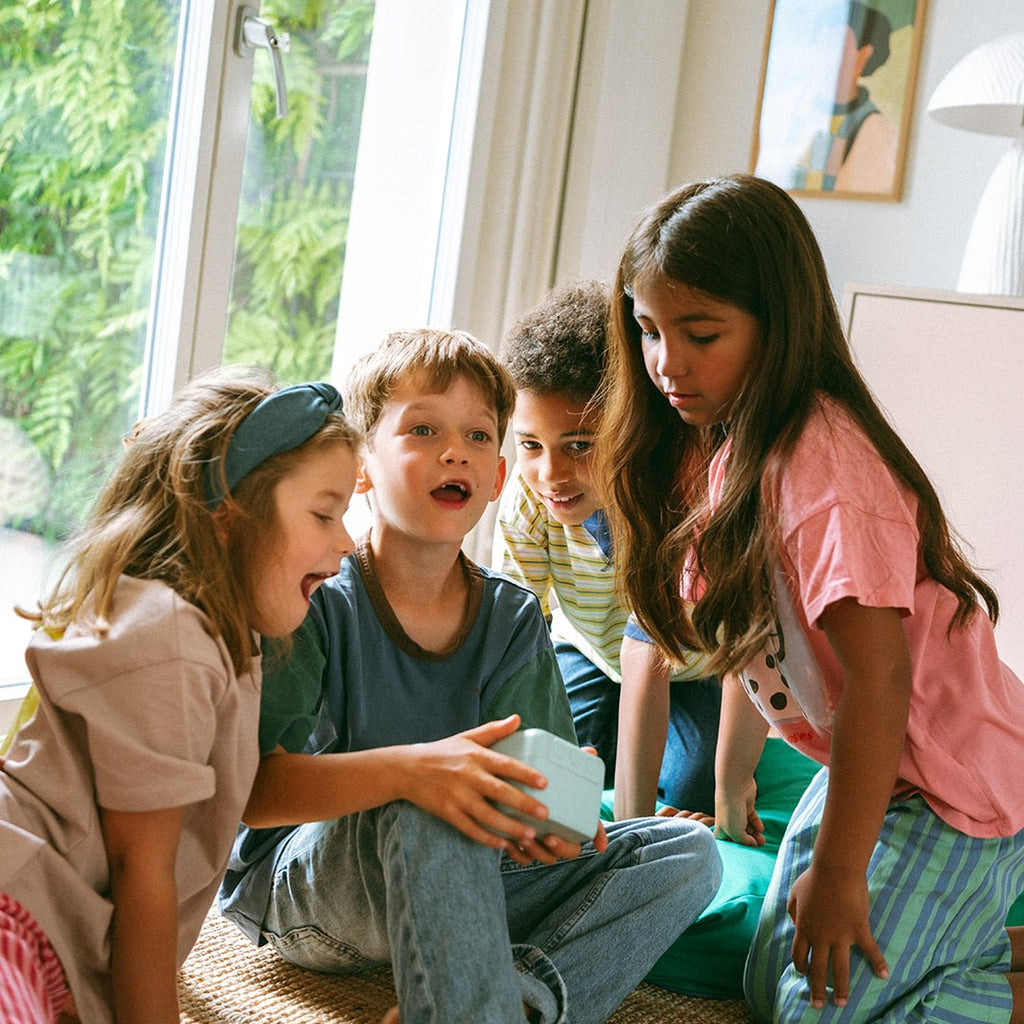 Four excited children sitting on a rug by a sunny window gathered round the green KIDYPRINTER AI