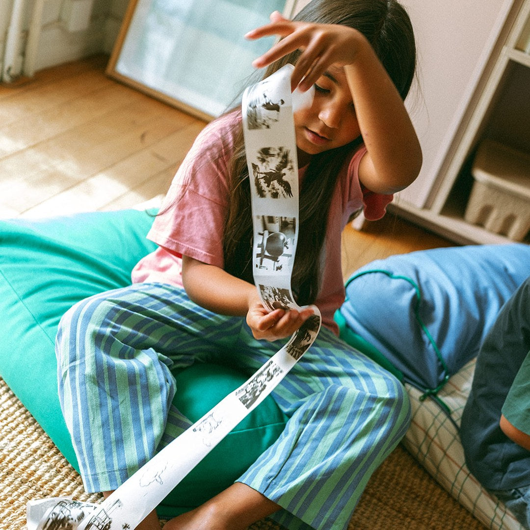 Girl in striped trousers sitting on a beanbag looking at a long strip of colouring prints from the KIDYPRINTER AI