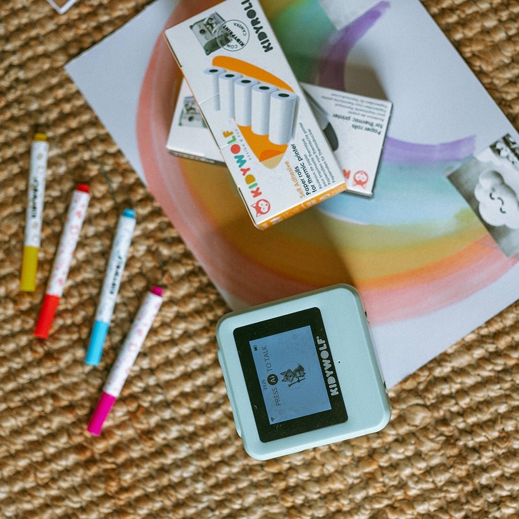 Green KIDYPRINTER AI on a jute rug with felt-tip markers, a box of paper refill rolls and a rainbow painting
