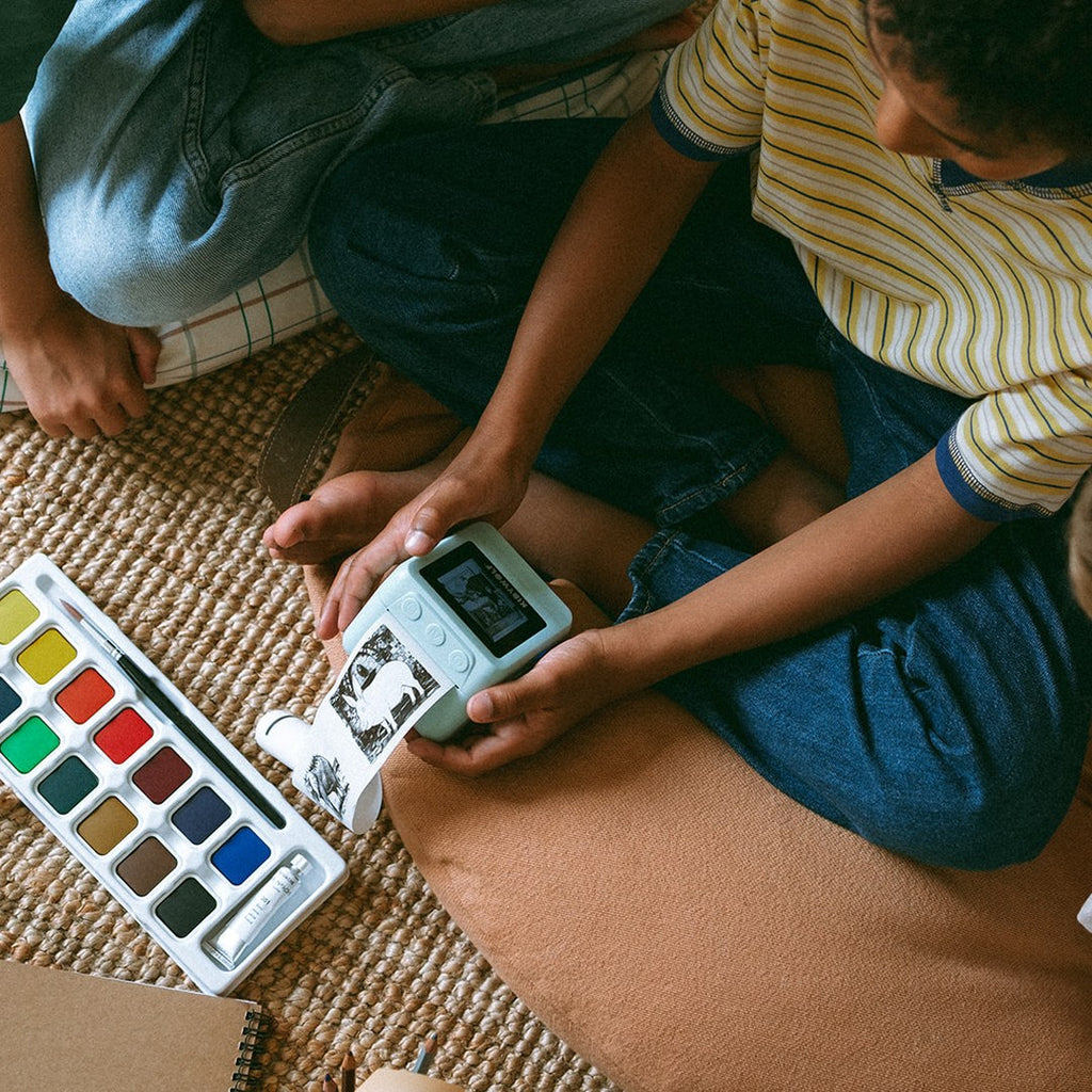 Child sitting on a rug printing with the green KIDYPRINTER AI next to a watercolour paint set and markers