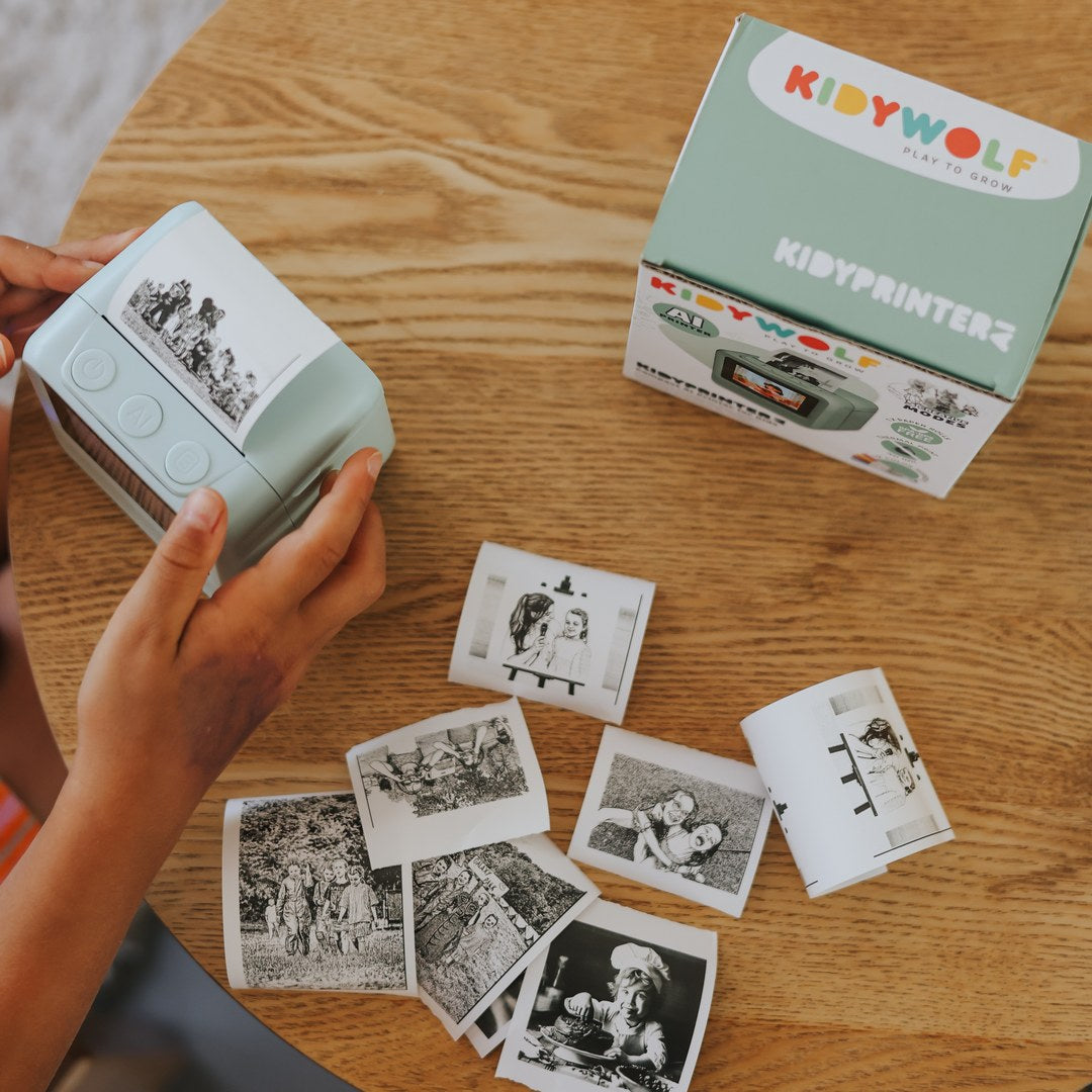 Hands holding the green KIDYPRINTER AI over a round wooden table scattered with prints, next to the KIDYPRINTER box