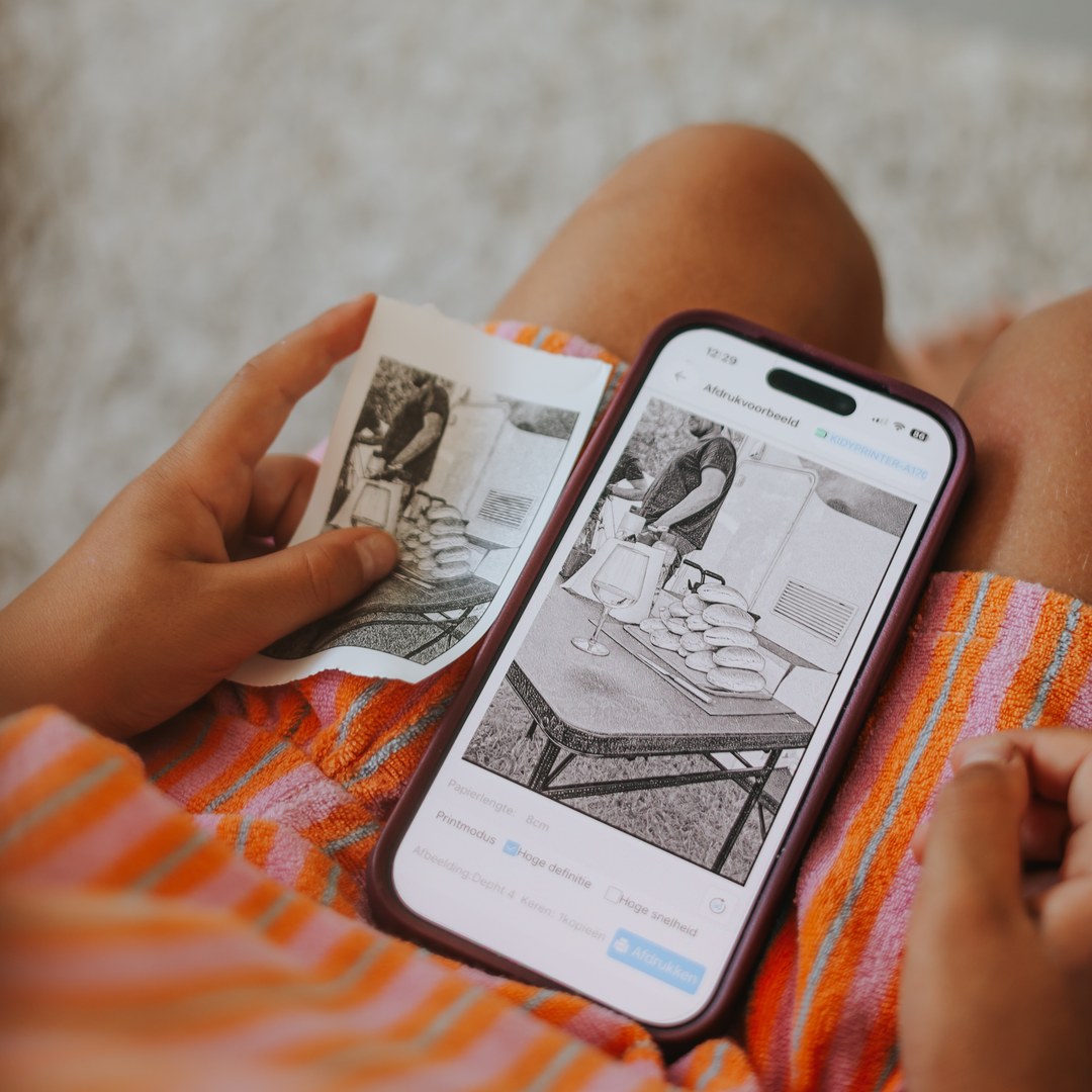 Child holding a small black and white KIDYPRINTER AI print beside a phone showing the same photo