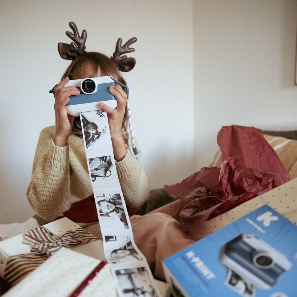 Child in reindeer antlers taking a photo with the blue K-PRINT as a long strip of printed photos falls onto Christmas presents and the K-PRINT box