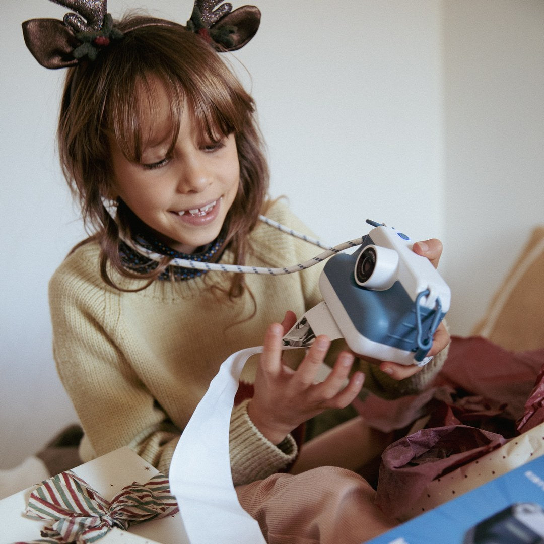 Smiling girl in reindeer antlers holding the blue K-PRINT camera as it prints among wrapped Christmas presents