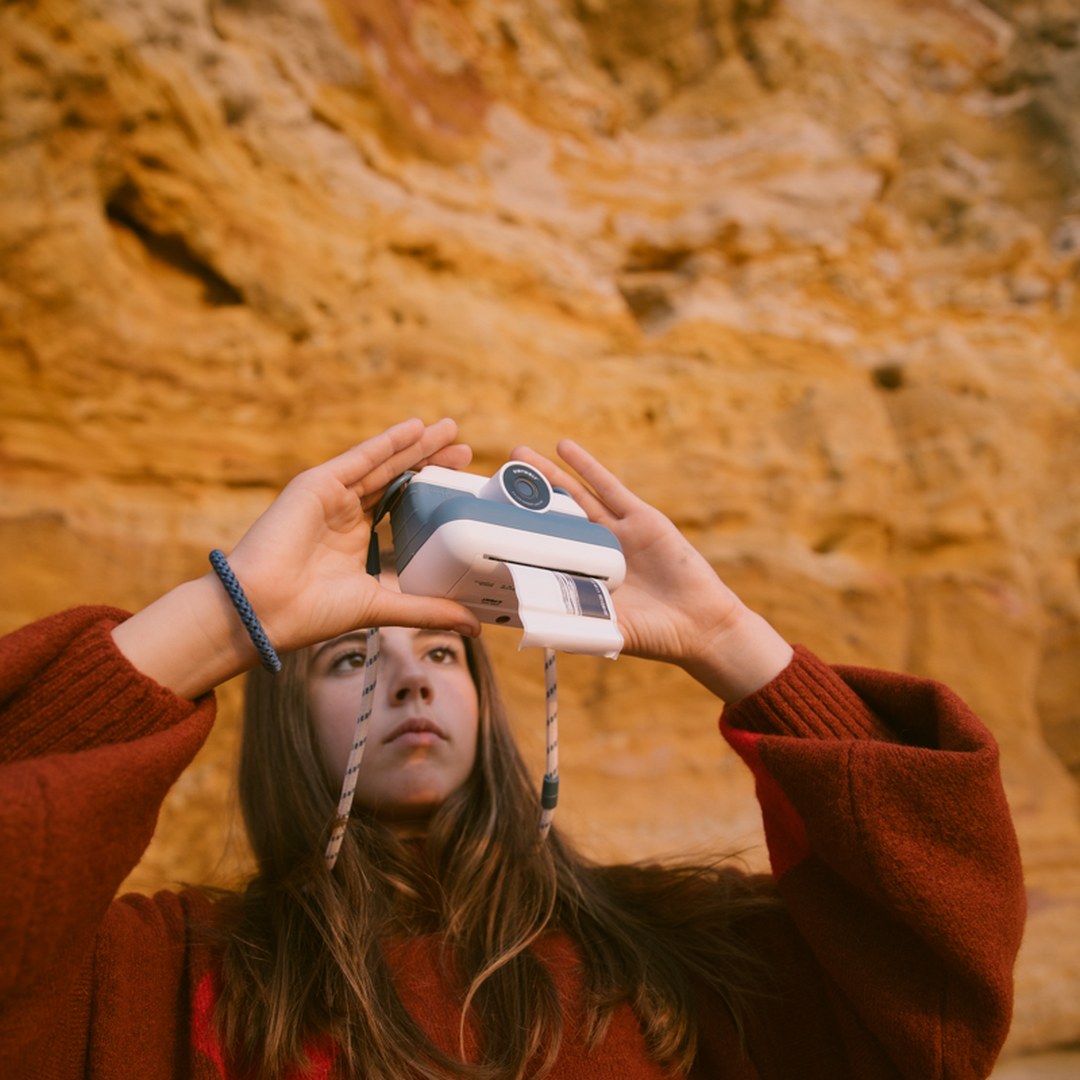 Girl in a red jumper holding the blue K-PRINT camera above their head in front of golden sandstone cliffs