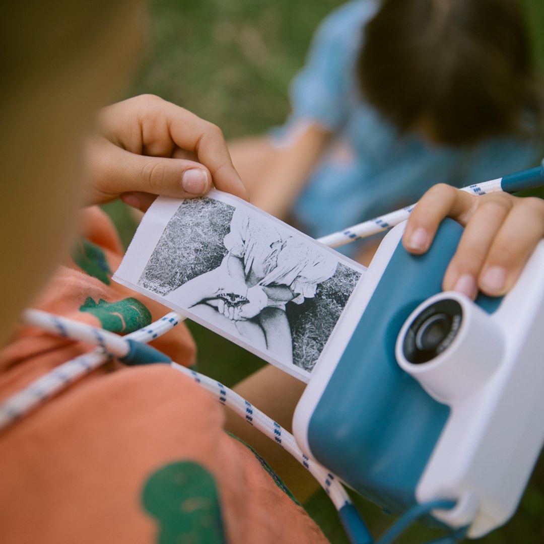 Close-up of a child's hands holding the blue K-PRINT camera and a printed black and white leaf photo