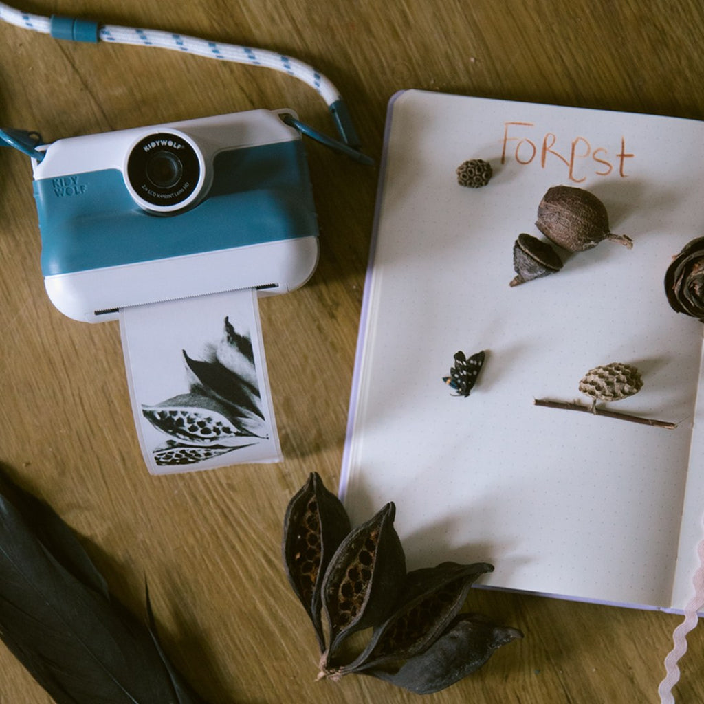 Front view of the blue K-PRINT camera printing a leaf photo next to a nature journal with acorns and seed pods