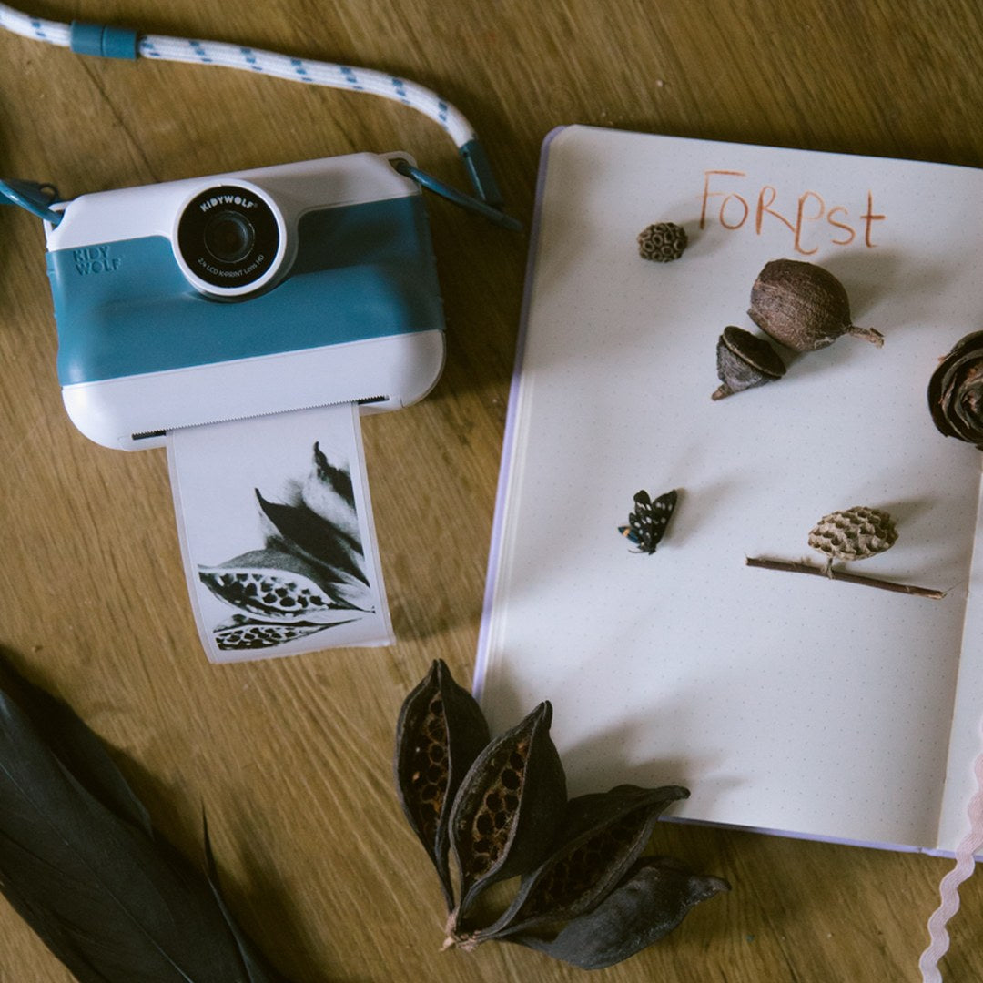 Front view of the blue K-PRINT camera printing a leaf photo next to a nature journal with acorns and seed pods