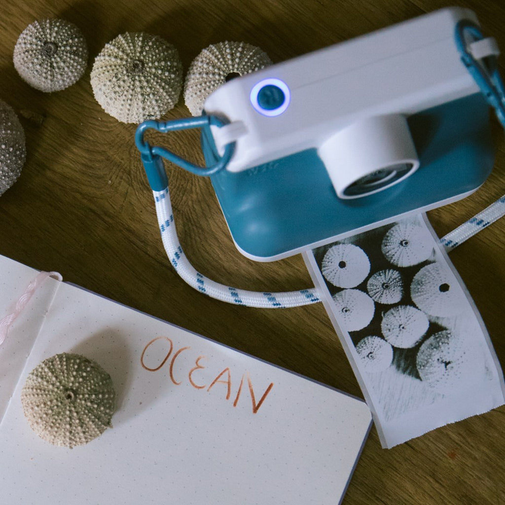 Blue K-PRINT camera with its rope strap printing a photo of sea urchin shells beside an OCEAN journal