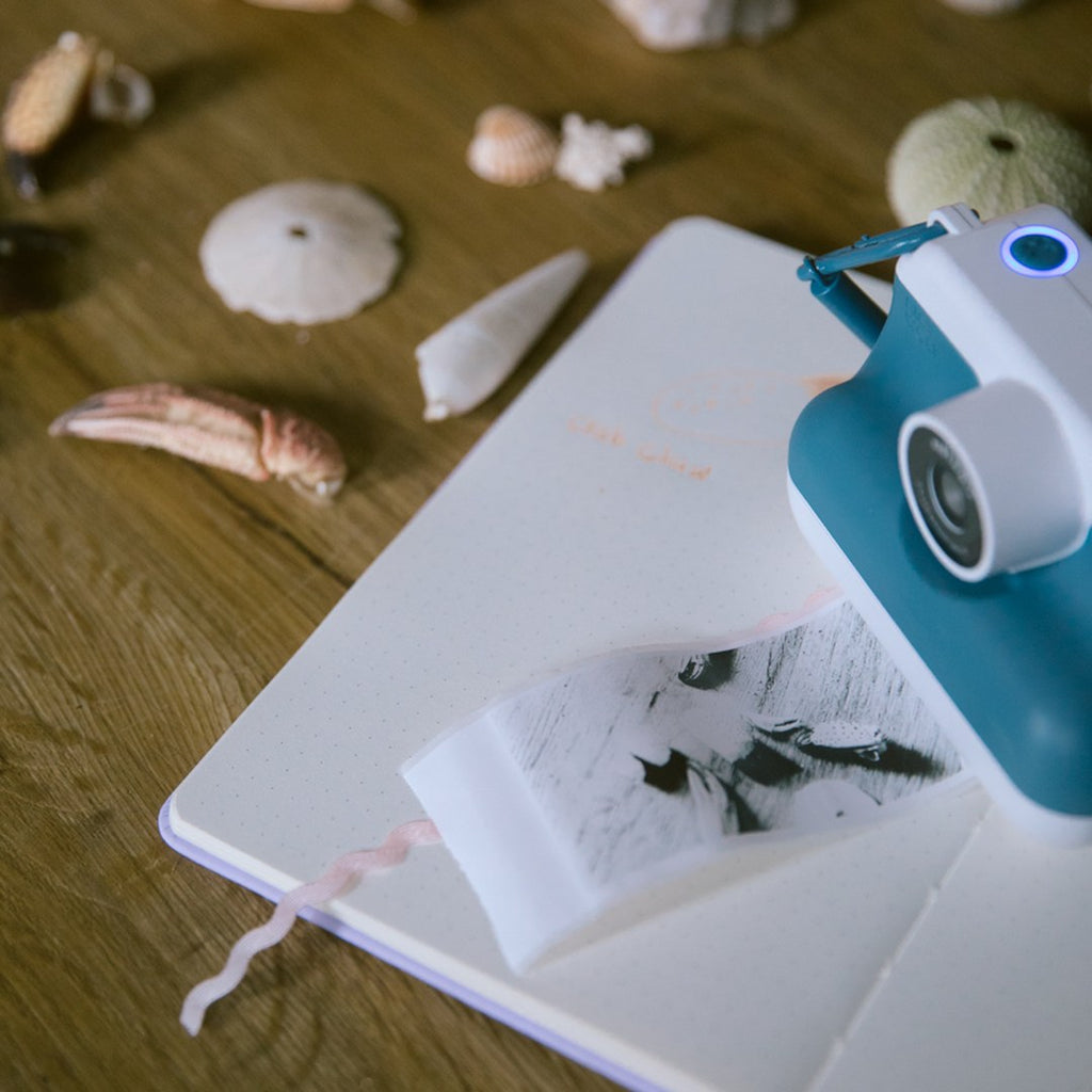 Blue K-PRINT camera on an open notebook printing a black and white photo with seashells scattered on the table