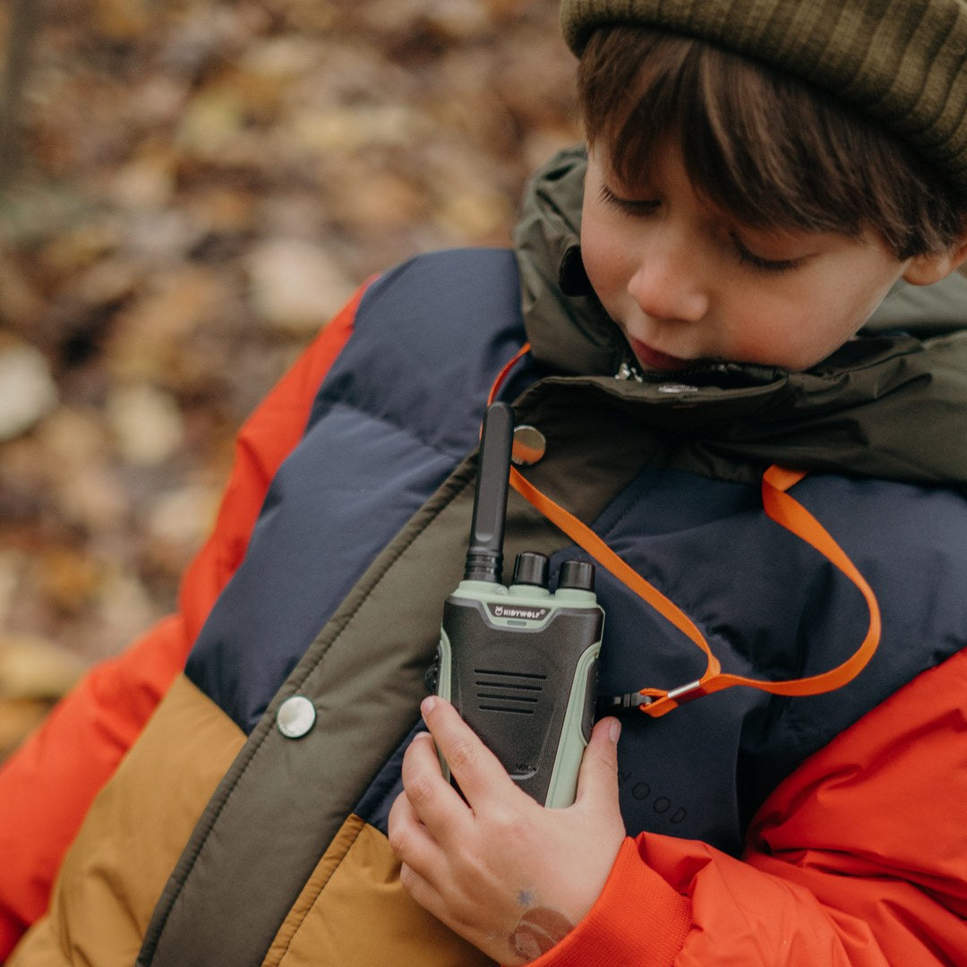 Boy in a woolly hat and puffer jacket holding the green KIDYTALK walkie-talkie on an orange lanyard in autumn woods