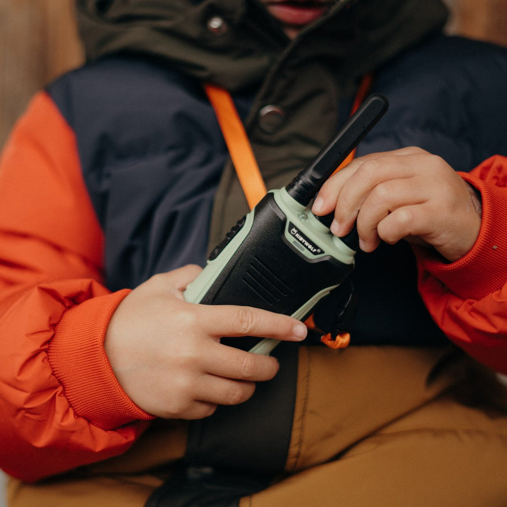 Close-up of a child in an orange and navy coat turning the dial on the green KIDYTALK walkie-talkie