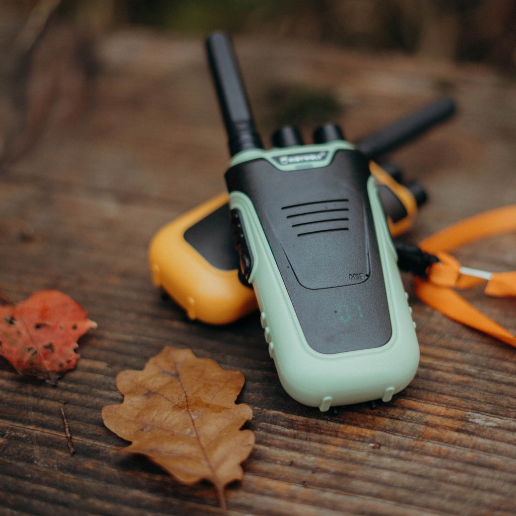 Green and orange KIDYTALK walkie-talkies lying on a wooden table with autumn leaves and an orange lanyard