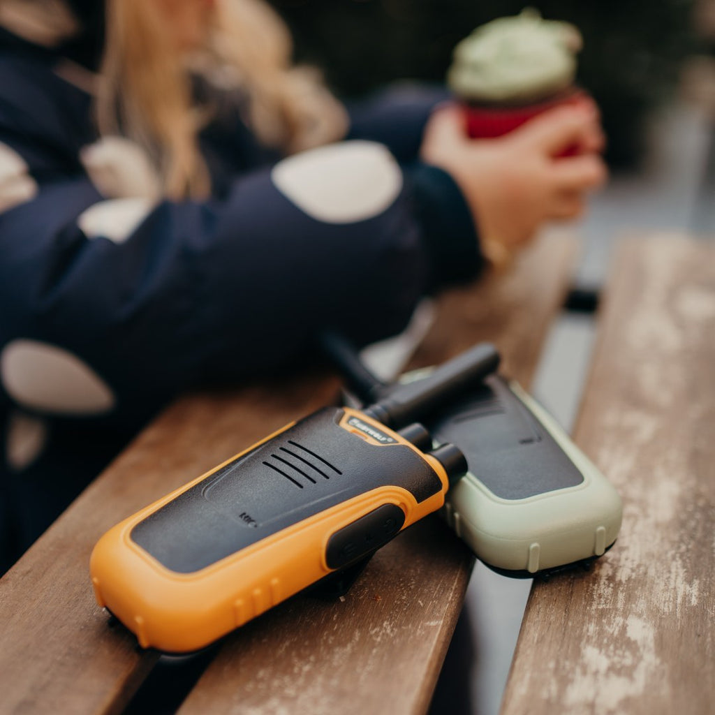 Orange and green KIDYTALK walkie-talkies on a wooden bench while a child in a red hat drinks hot chocolate