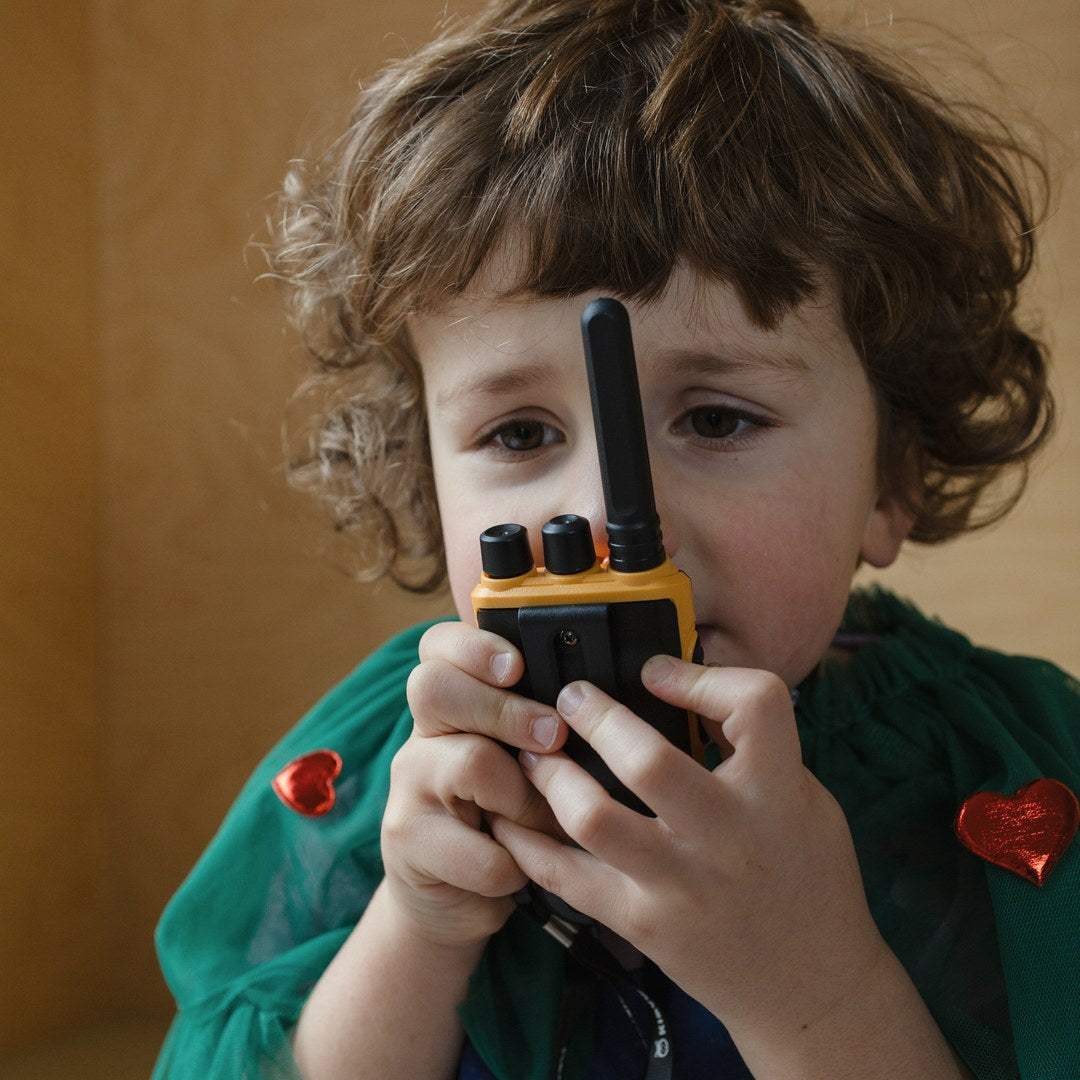 Curly-haired boy in a green cape talking into the orange KIDYTALK walkie-talkie